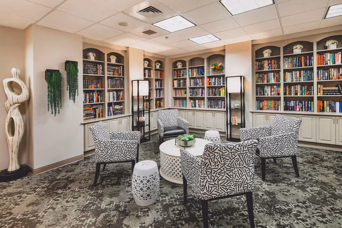 A cozy library room with built-in bookshelves filled with books along the walls. Four patterned armchairs are arranged around a round white coffee table with a small plant centerpiece. Two white decorative stools and two tall floor lamps are also present. The room has a patterned carpet and neutral-colored walls with some hanging greenery and a modern abstract sculpture in the corner.