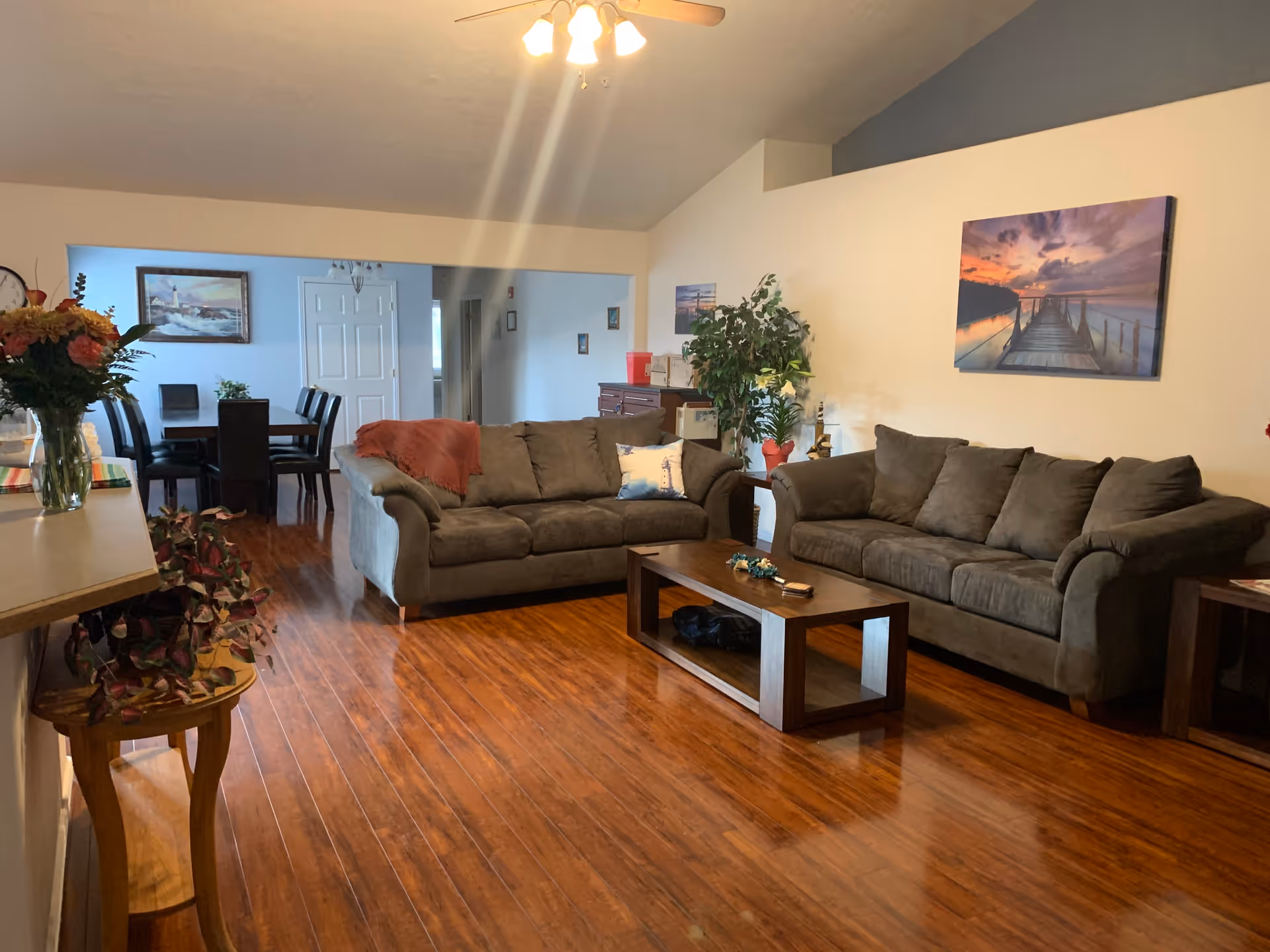 A cozy living room area with two gray sofas facing a wooden coffee table on a polished wooden floor. There are decorative pillows on the sofas, a plant in the corner, and framed pictures on the walls. In the background, a dining area with a table and chairs is visible, along with a hallway and a white door.