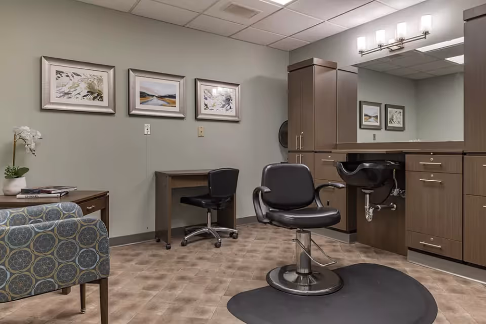 Interior of a salon room with a black salon chair in front of a sink and large mirror, wooden cabinets, a small desk with a black swivel chair, a patterned armchair, and three framed artworks on the wall.