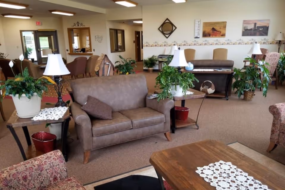 A spacious senior living facility common area with multiple seating options including brown leather sofas, patterned armchairs, and wooden tables adorned with white lace doilies and potted plants. The room has beige carpeting, wall decorations, and soft lighting from table lamps. There are large windows and doors in the background letting in natural light.