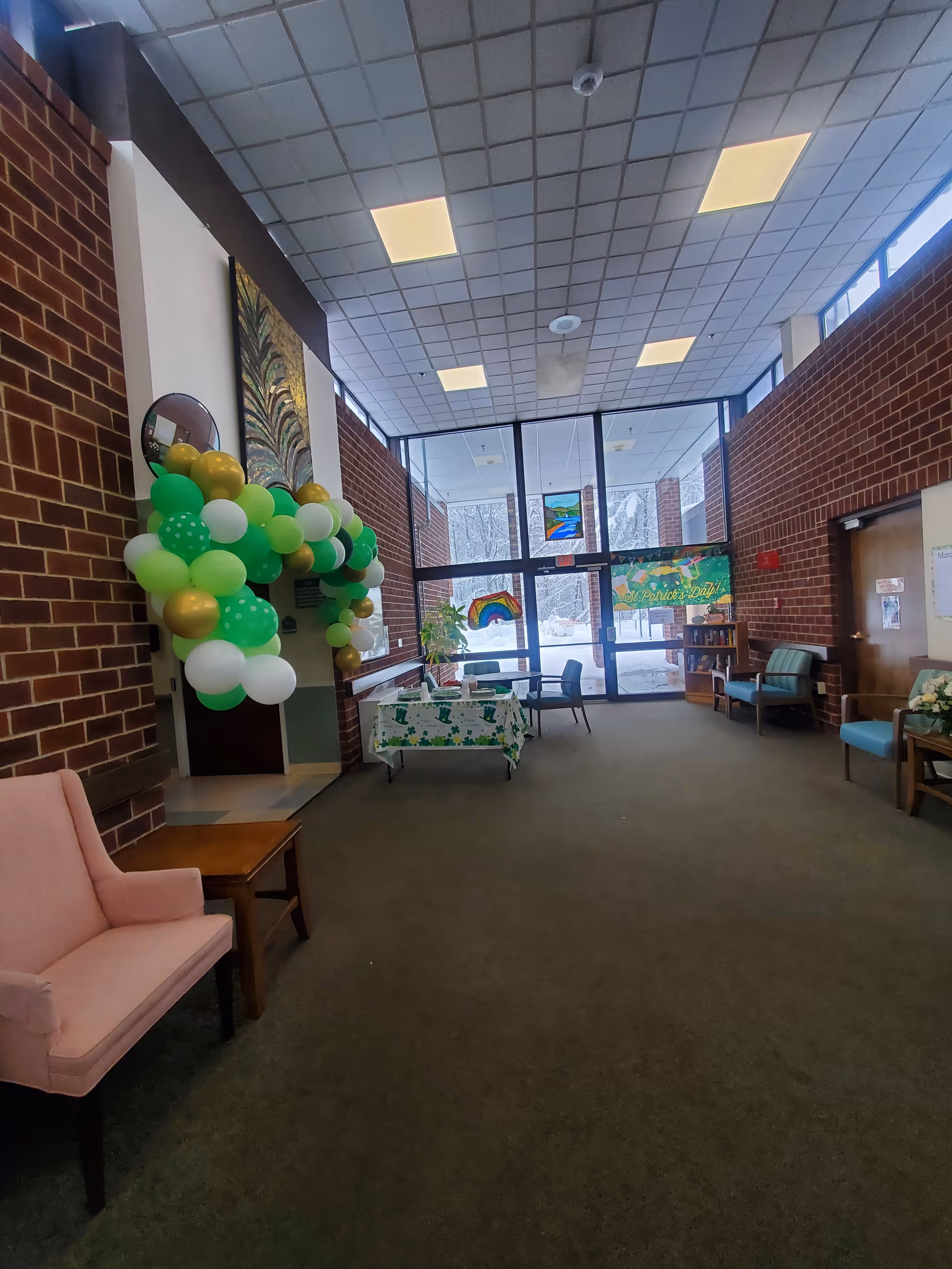 A spacious lobby area with brick walls and large windows letting in natural light. The room features a pink armchair and a wooden side table on the left, a table decorated with a green and white tablecloth and green, white, and gold balloons arranged in an arch. There are several chairs and a small bookshelf near the windows. Outside, snow is visible through the glass doors and windows.