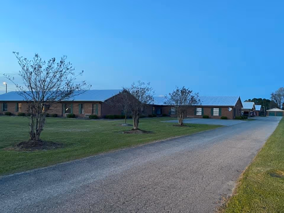Exterior view of a single-story brick building with a metal roof, surrounded by a grassy lawn and a few small trees, under a clear blue sky at dusk.