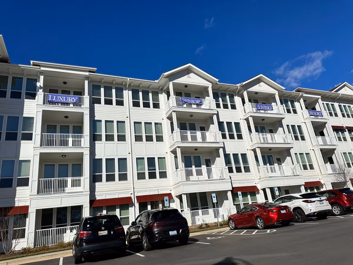 White multi-story senior living building with balconies, banners reading 'LUXURY SENIOR LIVING RESORT', and cars parked in front under a clear blue sky.