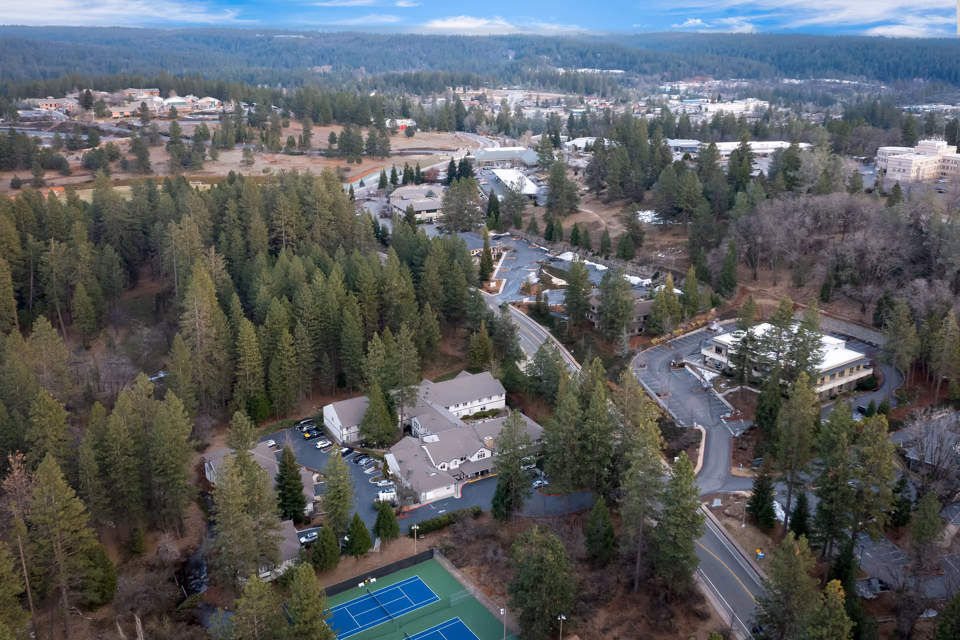 Aerial view of Cascades of Grass Valley facility surrounded by dense trees and greenery, with multiple buildings, parking areas, roads, and a tennis court visible under a partly cloudy sky.