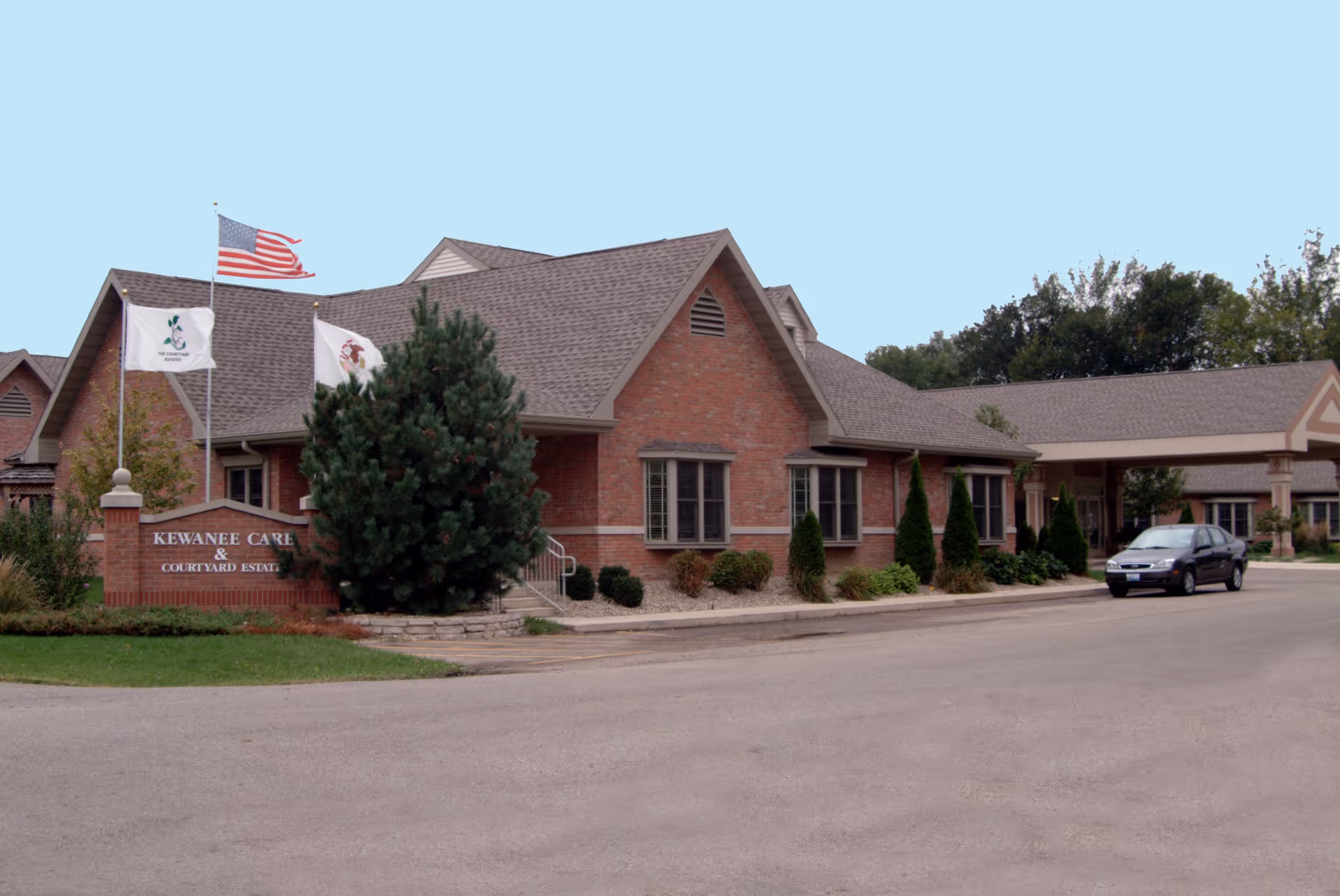 Brick one-story senior care facility entrance with flags, landscaping, and a sign reading 'Kewanee Care'.