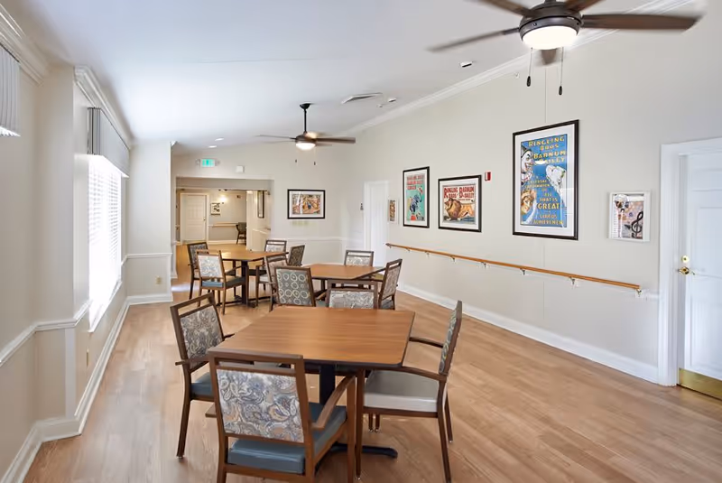 A bright dining room with wooden tables and chairs featuring patterned upholstery. The room has light-colored walls adorned with framed vintage circus posters. Ceiling fans with lights hang from the ceiling, and large windows on the left side allow natural light to fill the space. A wooden handrail runs along the right wall.