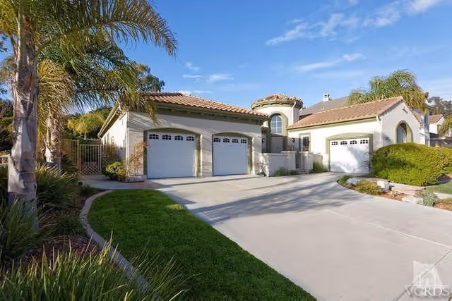 Exterior view of a residential building with a driveway leading to three white garage doors. The building has a light-colored stucco finish with a red tile roof and is surrounded by palm trees and well-maintained landscaping under a clear blue sky.