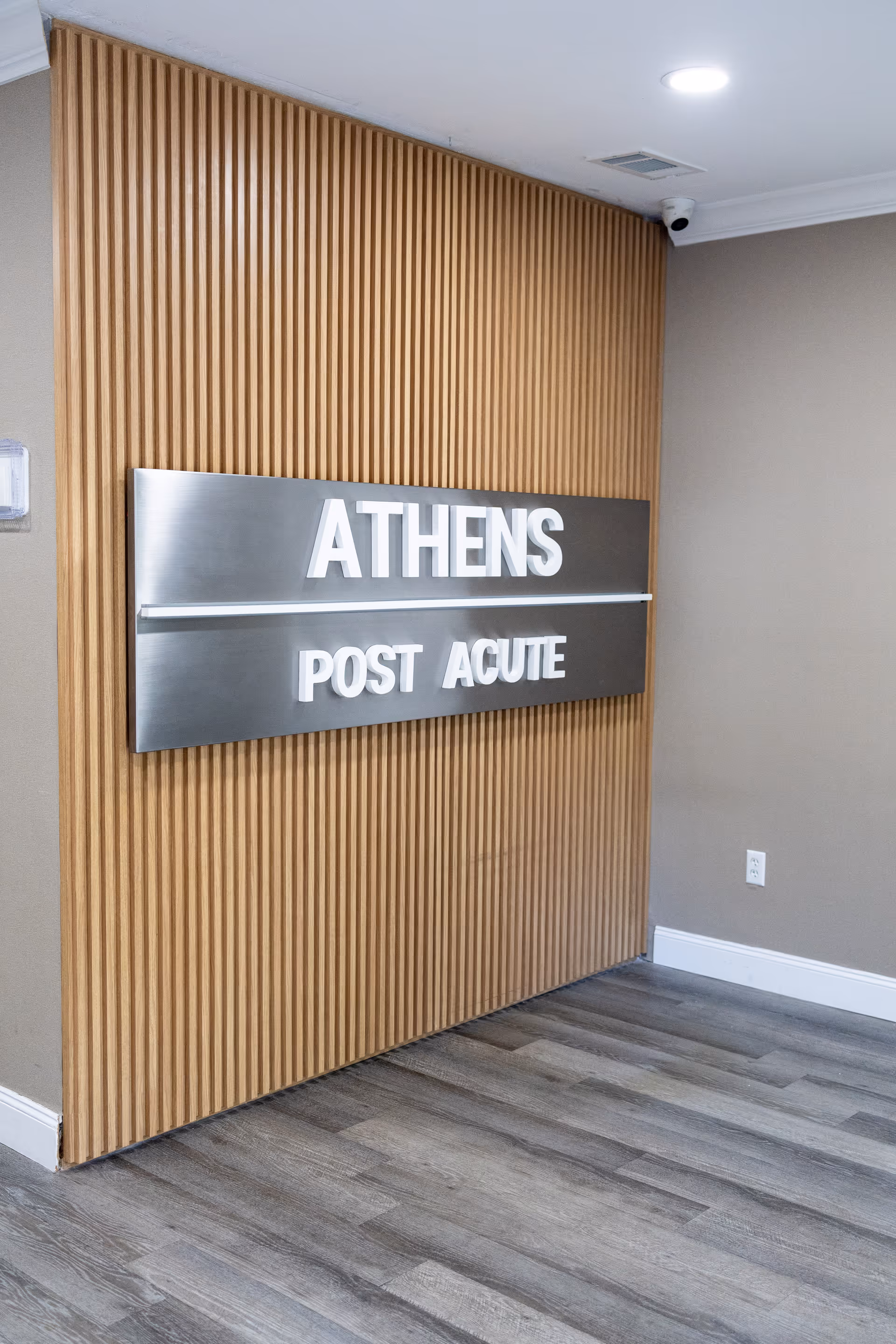 Interior corner of a facility with a wooden slat wall panel and a metallic sign mounted on it that reads 'ATHENS POST ACUTE'. The floor has gray wood-like planks, and the walls are painted beige with white baseboards. A security camera is mounted on the ceiling.