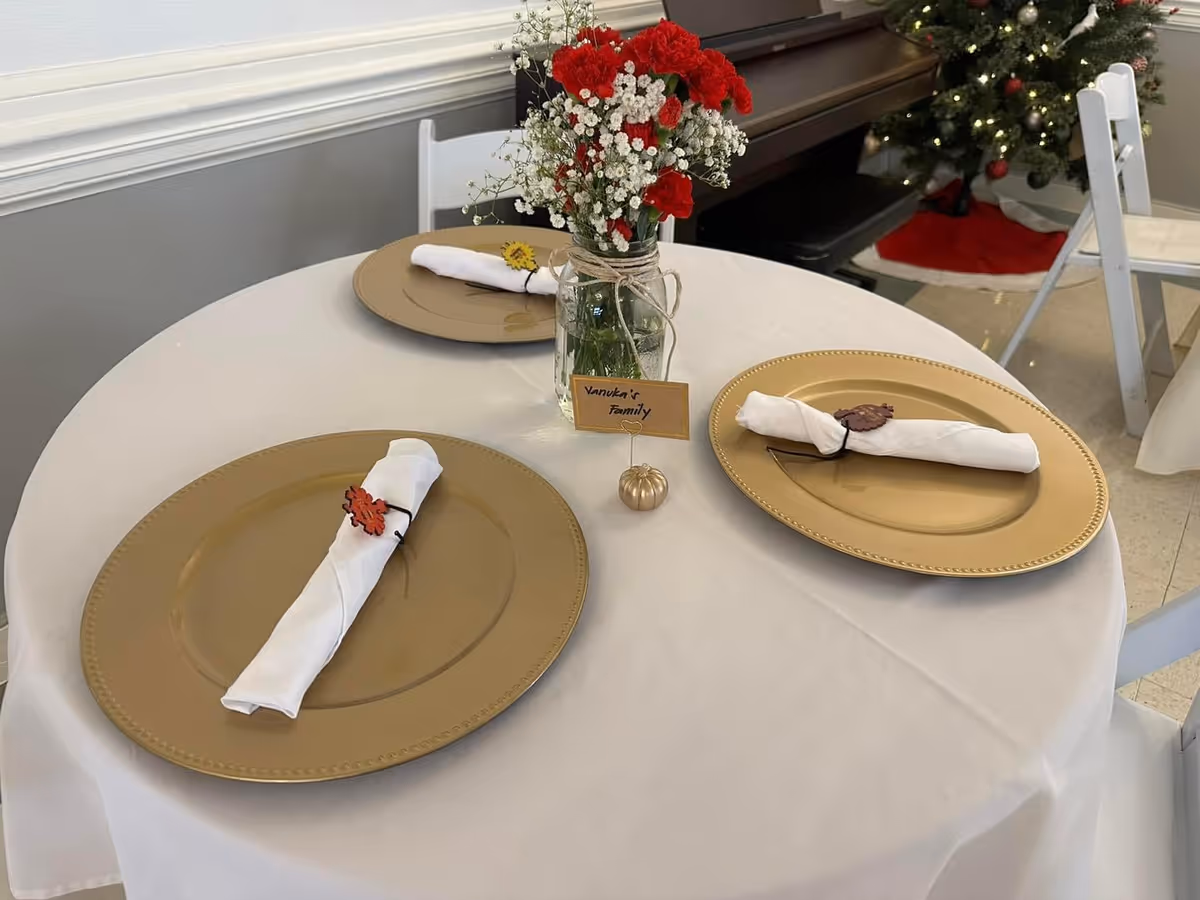 A round table set for three with gold charger plates and white rolled napkins secured with decorative napkin rings. A small vase with red and white flowers is placed in the center of the table with a card labeled 'Vanuka's Family'. In the background, a decorated Christmas tree and a piano are visible.