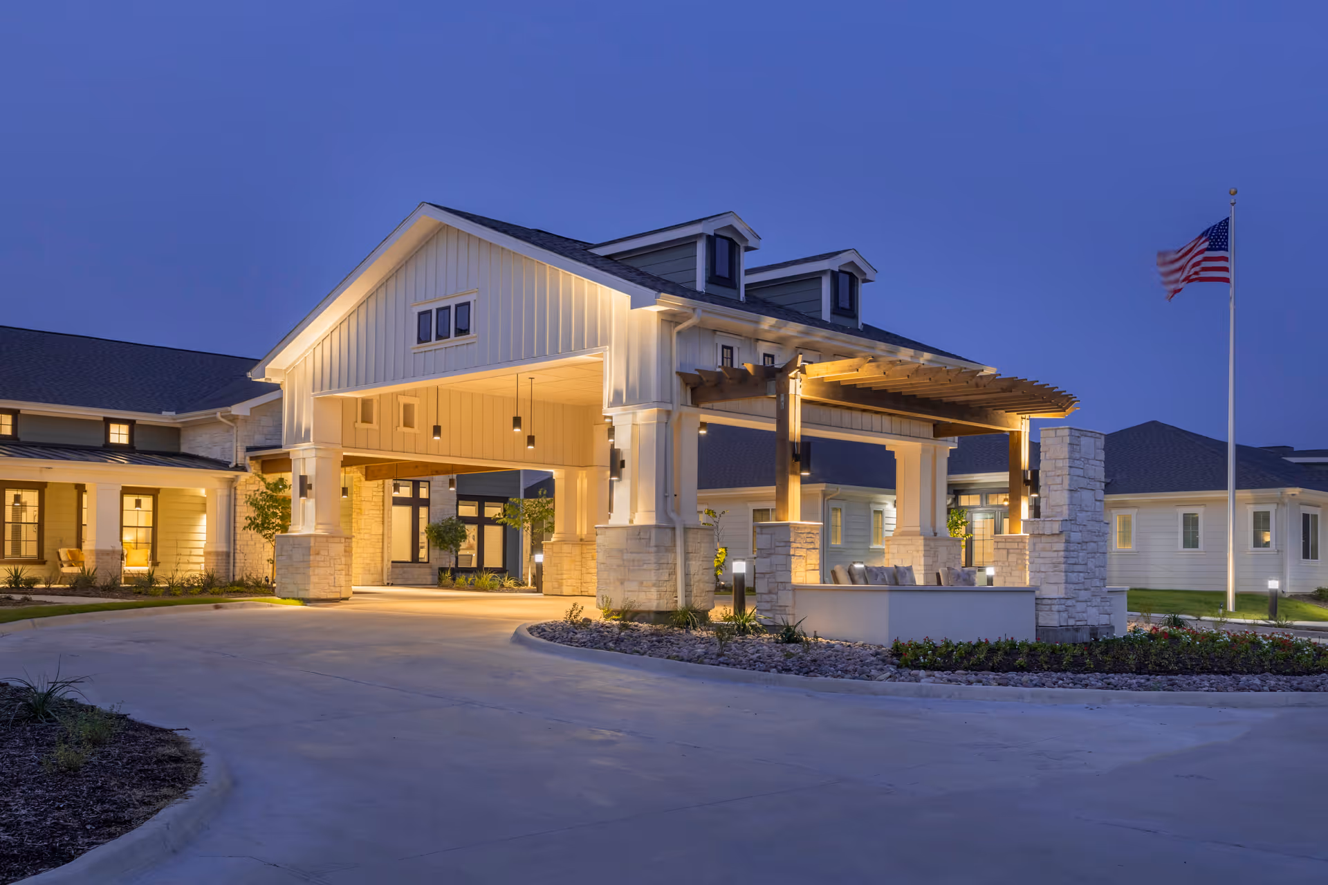Exterior view of The Preserve at Spring Creek facility at dusk, showing a well-lit entrance with a covered driveway, stone pillars, and an American flag on a flagpole to the right.