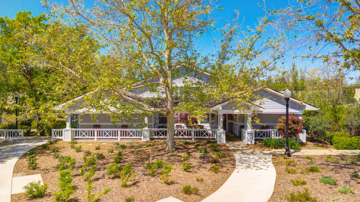 Front exterior of a single-story community building with a wraparound porch, tree in the foreground, and landscaped walkways.