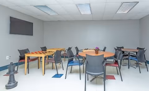 Communal activity room with round and rectangular tables, multiple chairs, a wall-mounted TV, and oversized chess pieces on the floor.