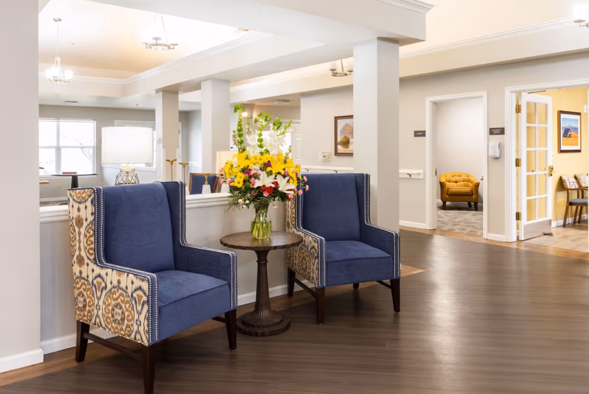 A bright senior living lobby with two blue patterned armchairs flanking a round table topped with a vase of flowers.