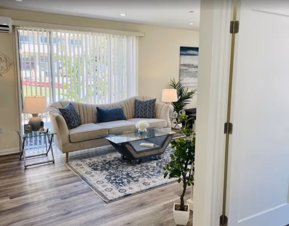 Bright living room with a beige sofa, glass coffee table on a patterned rug, lamps, and a potted plant in front of vertical blinds.