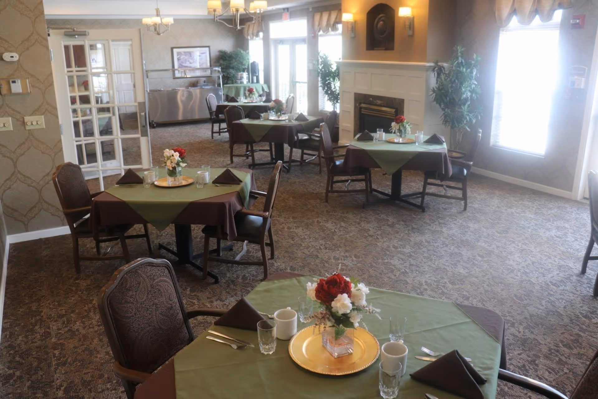 A dining room with several tables covered in green and brown tablecloths, each set with glasses, mugs, silverware, and floral centerpieces. The room has carpeted floors, a fireplace, large windows with blinds, and a serving area in the background.