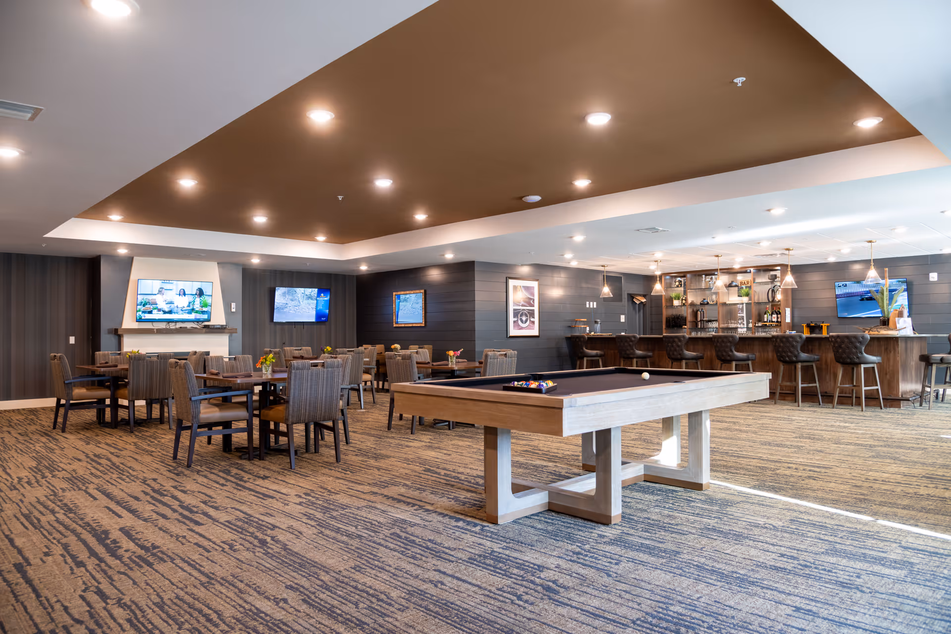 A spacious common area in The Goldton at Venice featuring a pool table in the foreground, multiple tables and chairs arranged for seating, a bar with high stools, and several wall-mounted televisions. The room has a modern design with recessed lighting and carpeted flooring.