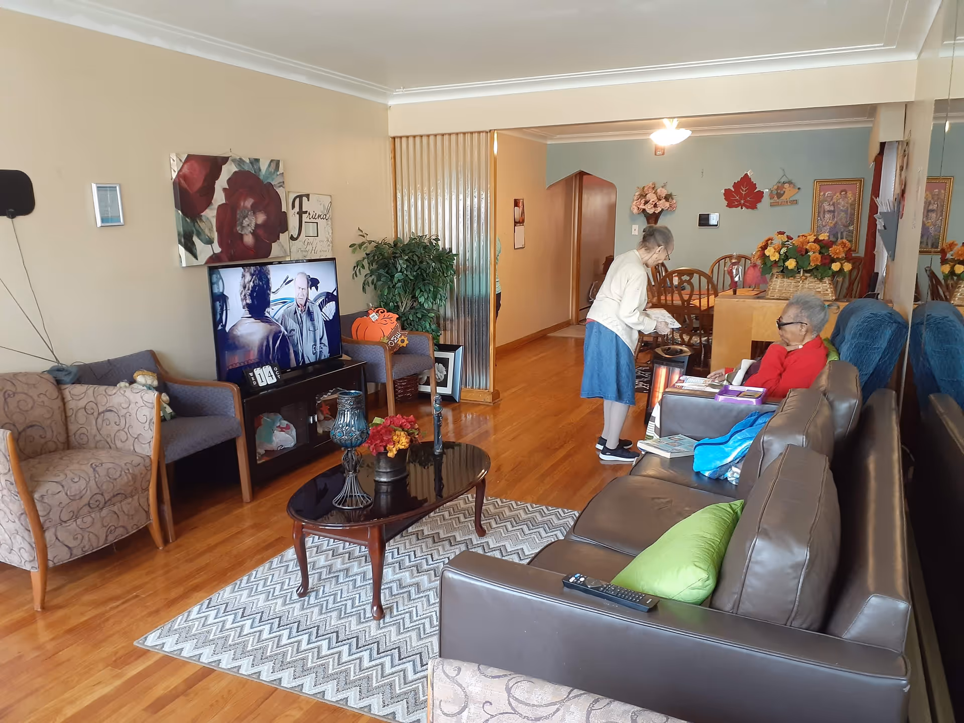 A cozy living room in an assisted living facility with wooden floors, a brown leather sofa with green pillows, a patterned armchair, and a TV showing a scene from a movie. Two elderly women are present, one standing and the other sitting on the sofa. The room is decorated with floral artwork, a potted plant, and a dining area with a wooden table and chairs in the background.
