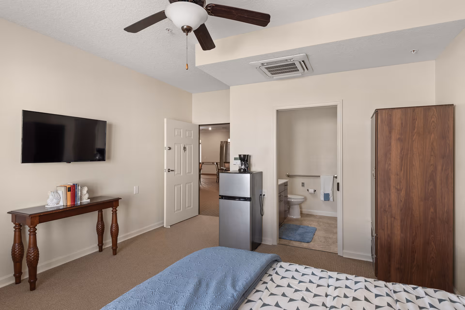 Interior view of a senior living bedroom featuring a bed with patterned bedding and a blue blanket, a wooden wardrobe, a small refrigerator with a coffee maker on top, a wall-mounted flat-screen TV above a wooden table with books, and an open door leading to a bathroom with a toilet and blue bath mats.