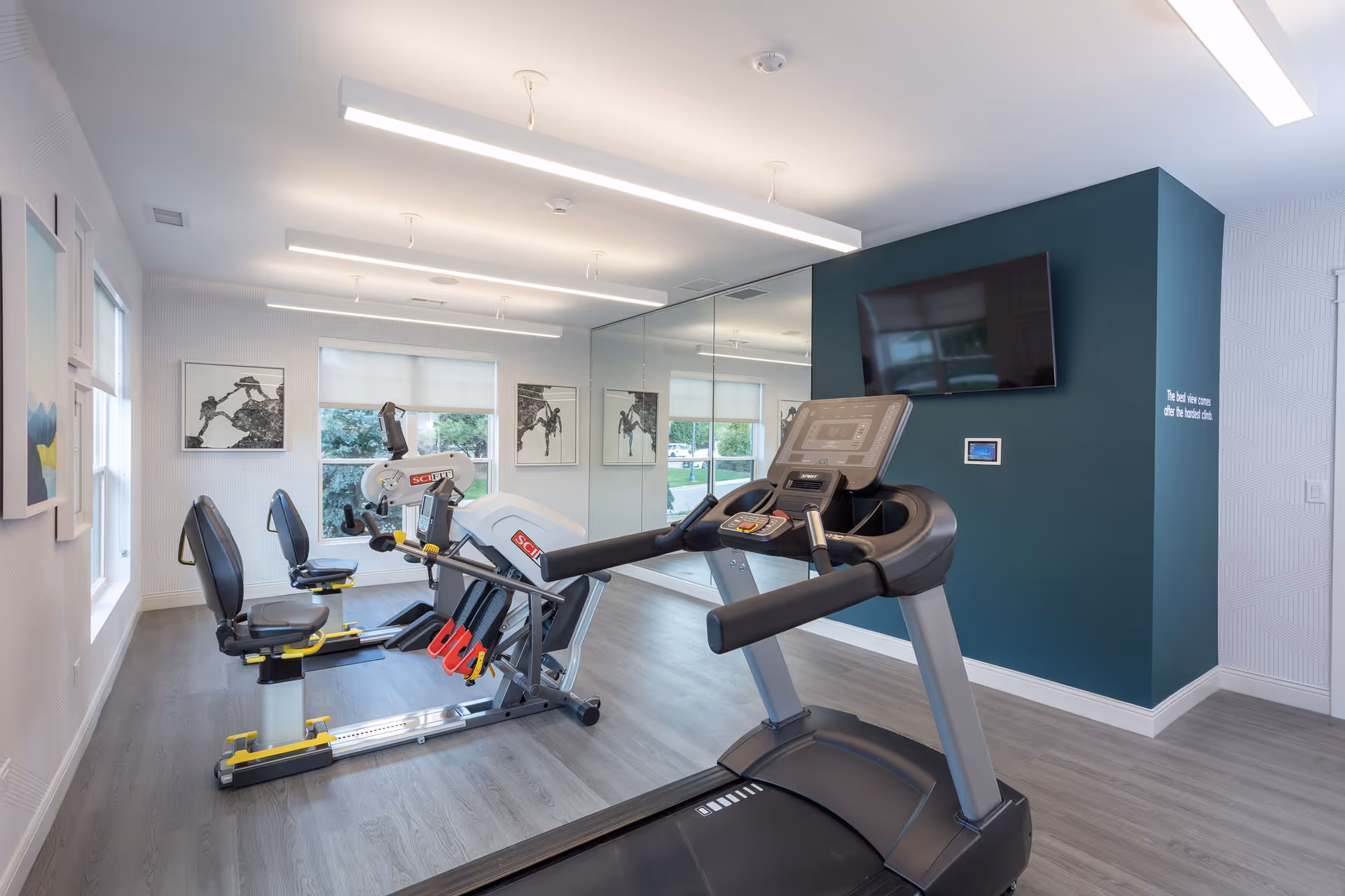 A bright and modern fitness room with exercise equipment including a treadmill and two recumbent bikes. The room has large windows, a wall-mounted TV, and a large mirror covering one wall. The walls are painted white with one accent wall in teal, and the floor is light wood.