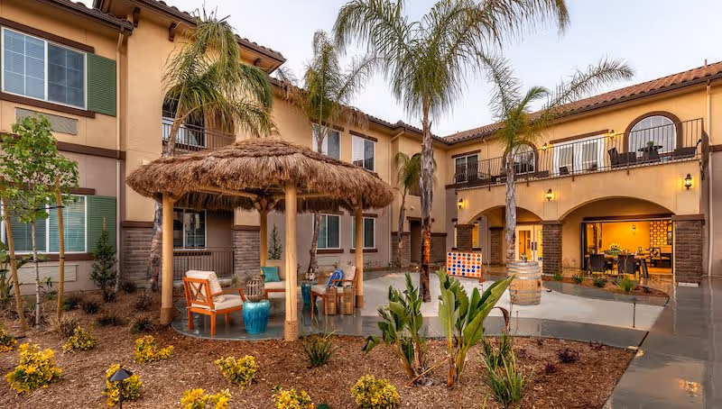 Outdoor courtyard area of a senior living facility with a thatched-roof gazebo, seating arrangements including chairs and a small table, palm trees, and landscaped garden beds. The building surrounding the courtyard has two stories with balconies and arched doorways, warmly lit from the inside.