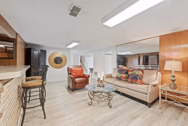A cozy living room with light wood flooring, a beige sofa with colorful pillows, a red leather armchair, a glass coffee table with decorative candles, a side table with a lamp, and a bar counter with two stools. The room has a large mirror on one wall and a round decorative wall piece.