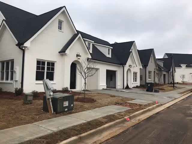 Row of newly constructed white cottages with black roofs along a paved sidewalk and street under a cloudy sky. The area appears to be part of a residential or senior living community with minimal landscaping and some utility boxes visible.