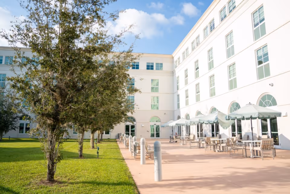 Outdoor patio area of a senior living facility with tables, chairs, and umbrellas set up along the side of a white multi-story building. There are trees and a grassy area adjacent to the patio under a partly cloudy sky.