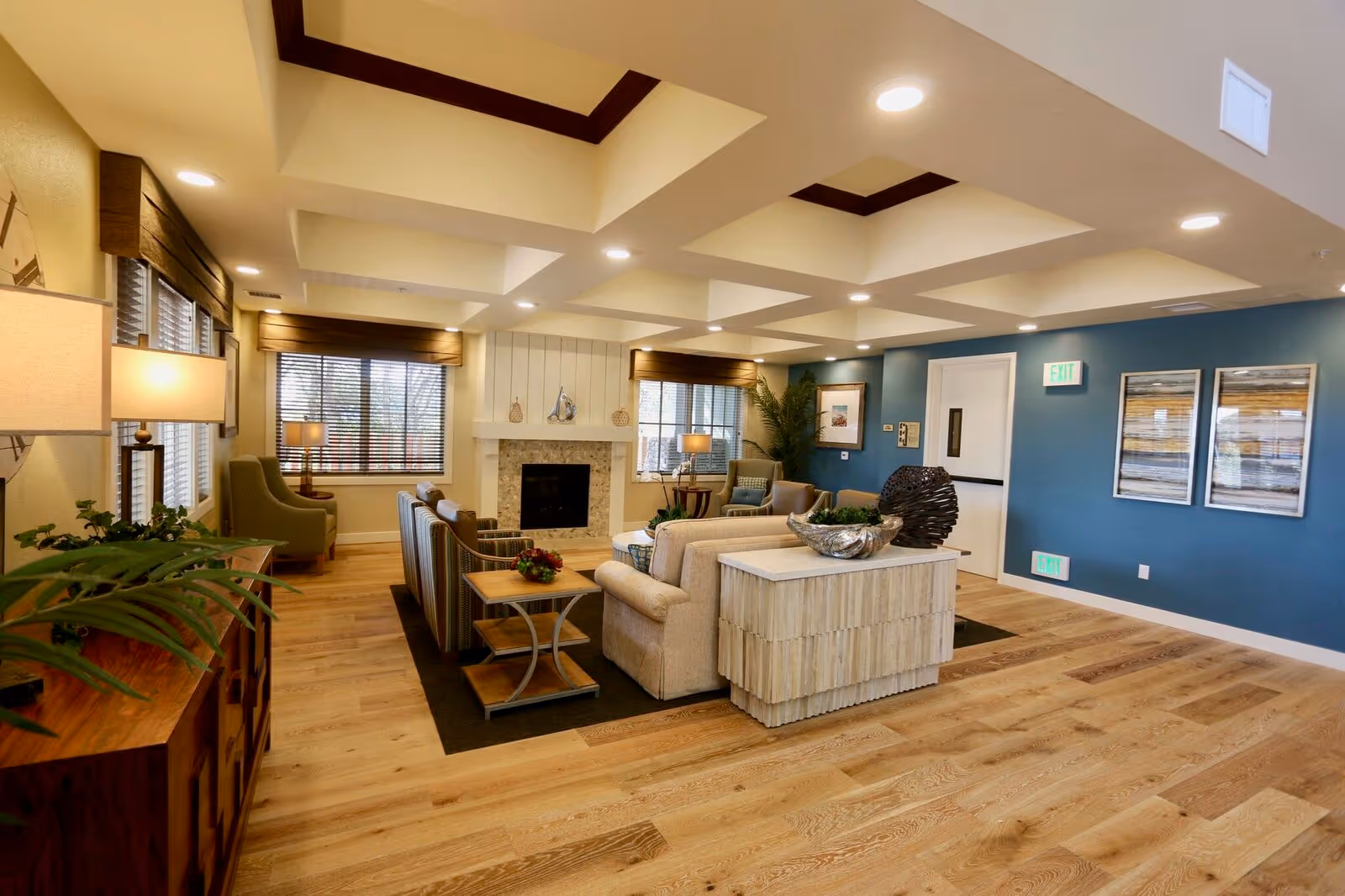 A spacious and well-lit living room area in a senior living facility with wooden flooring, beige and green armchairs, a beige sofa, a fireplace with decorative items on the mantel, large windows with blinds, modern ceiling lights, and blue accent walls with framed artwork.
