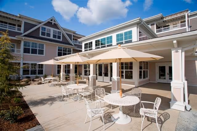Outdoor patio area of The Stafford Retirement Community with white metal tables and chairs, beige umbrellas providing shade, and a multi-story building with large windows in the background under a partly cloudy blue sky.