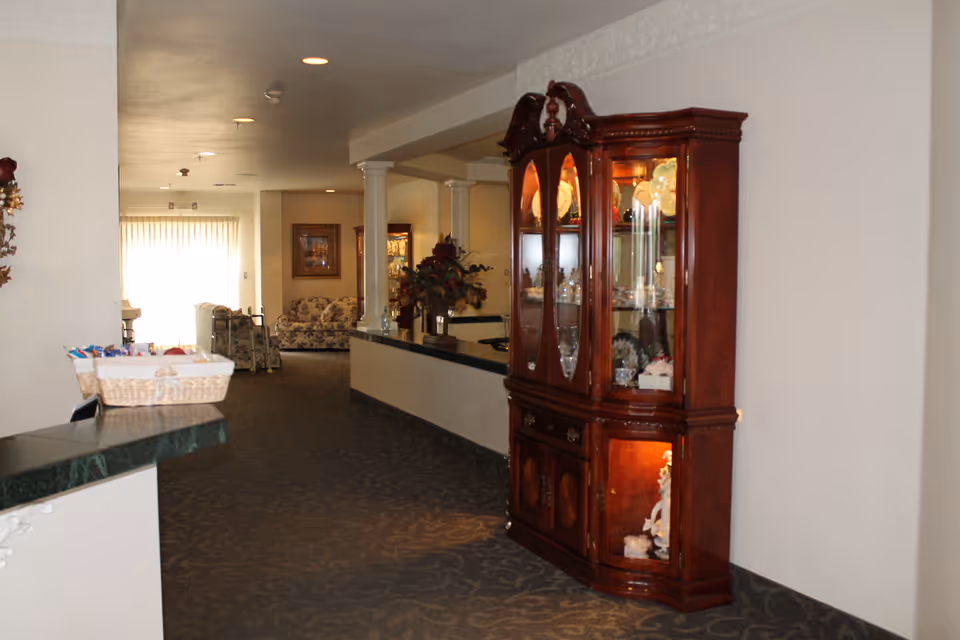 Interior hallway of a senior living facility with a dark carpeted floor, white walls, and ceiling lights. On the right side, there is a wooden display cabinet with glass doors showcasing decorative items. In the background, there are white columns, floral arrangements, and a seating area with floral-patterned sofas near a window with sheer curtains.