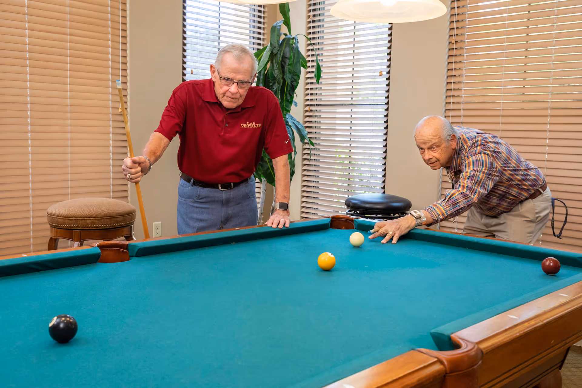 Two elderly men playing pool in a well-lit room with large windows covered by blinds. One man is holding a pool cue and wearing a red polo shirt with the text 'Las Ventanas,' while the other is aiming to take a shot. There is a plant and a stool in the background.