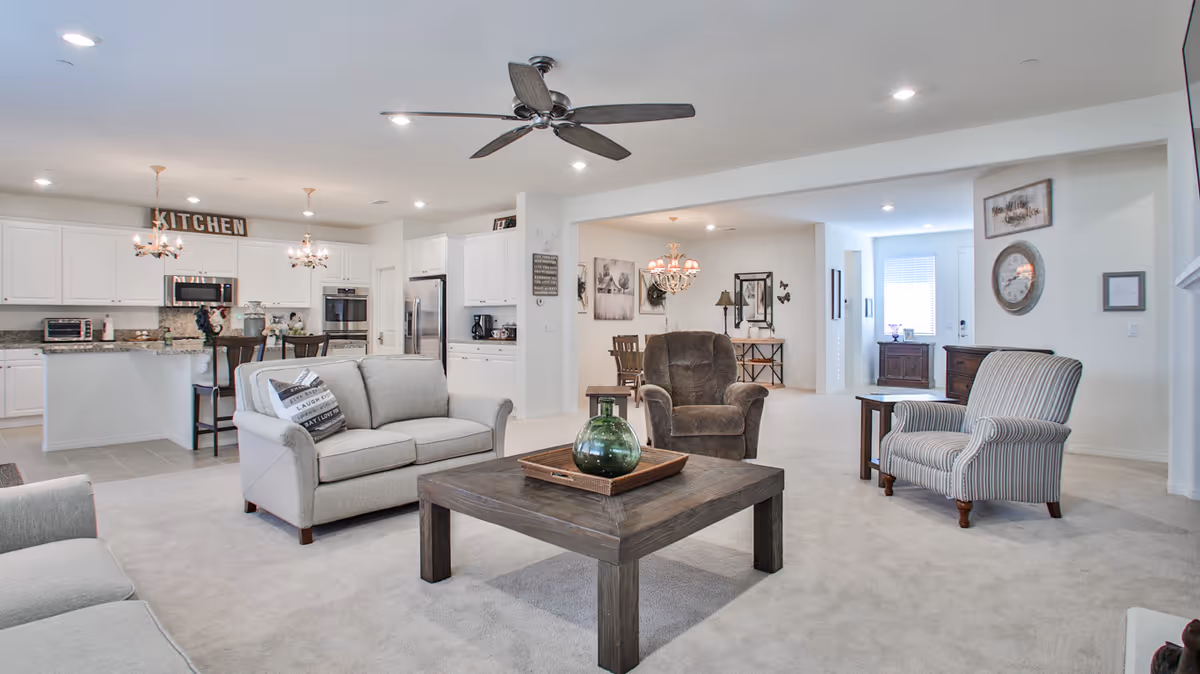 A spacious and bright living area featuring a beige carpet, a gray loveseat with decorative pillows, a brown armchair, and a striped armchair surrounding a wooden coffee table with a green vase. The open floor plan includes a white kitchen with granite countertops, stainless steel appliances, and a sign that reads 'KITCHEN' above the cabinets. A ceiling fan is mounted above the living area, and a dining area with a chandelier is visible in the background.