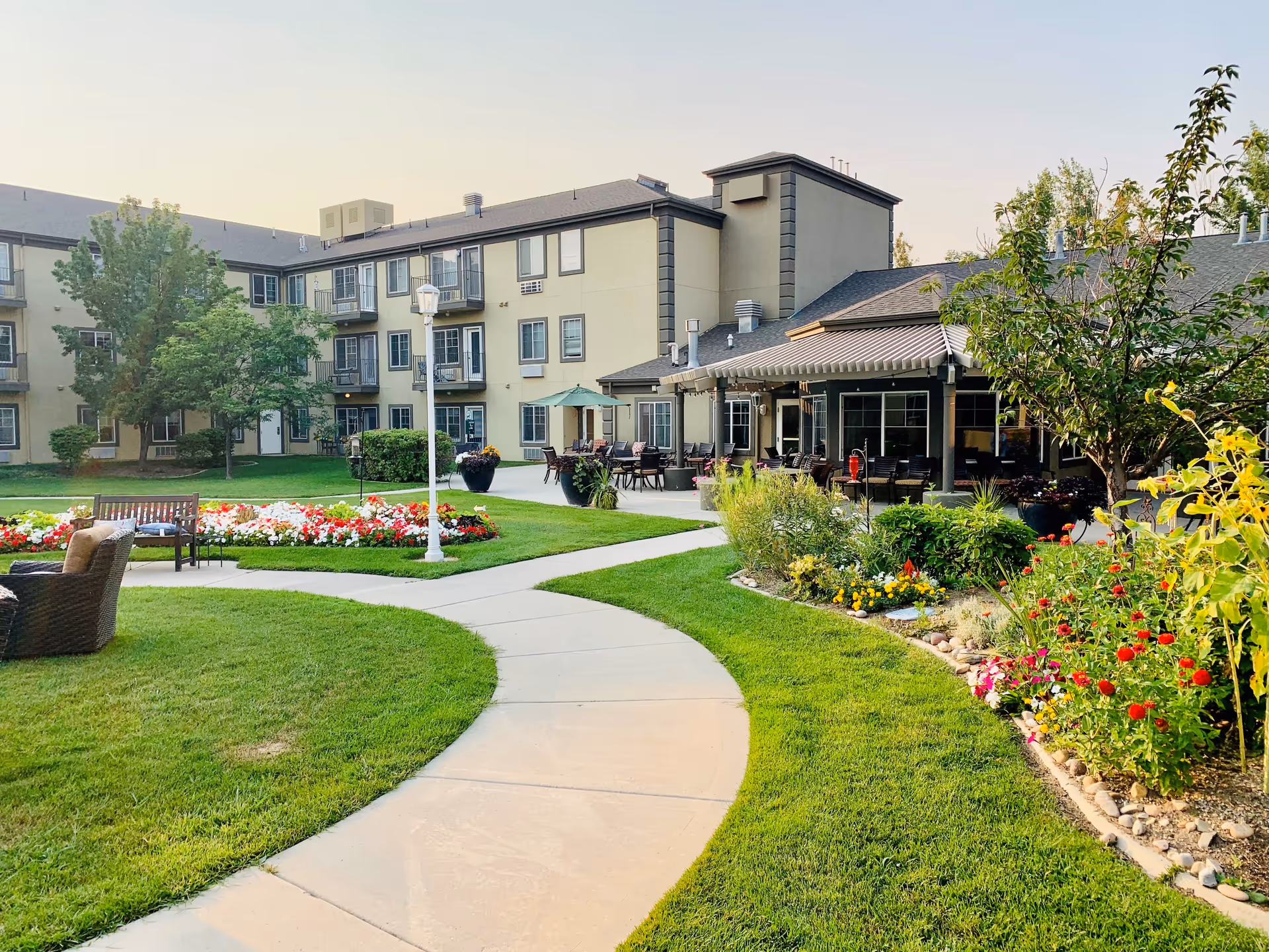 Outdoor view of Cedarwood at Sandy senior living facility showing a curved concrete pathway surrounded by green grass, colorful flower beds, benches, and patio seating areas. The multi-story building with balconies and large windows is visible in the background under a clear sky.