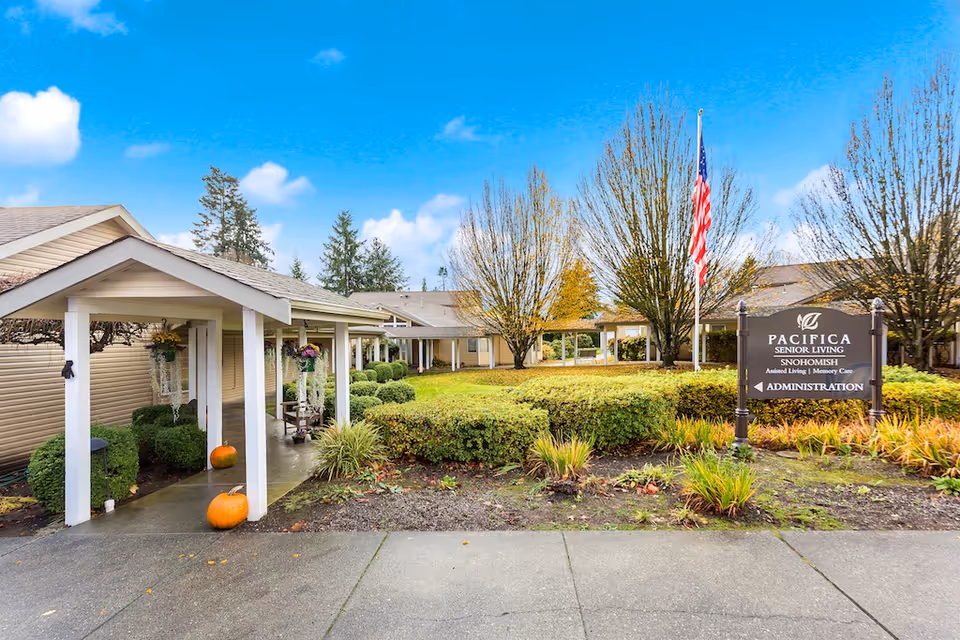 Front entrance of a senior living community with a covered walkway, landscaped courtyard, American flag, and a Pacifica Senior Living sign.