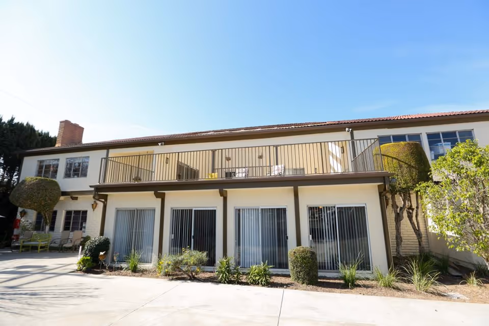 Exterior view of a two-story assisted living facility building with a balcony on the upper floor and large windows with vertical blinds on the ground floor. There are neatly trimmed bushes and plants along the building, and the sky is clear and blue.