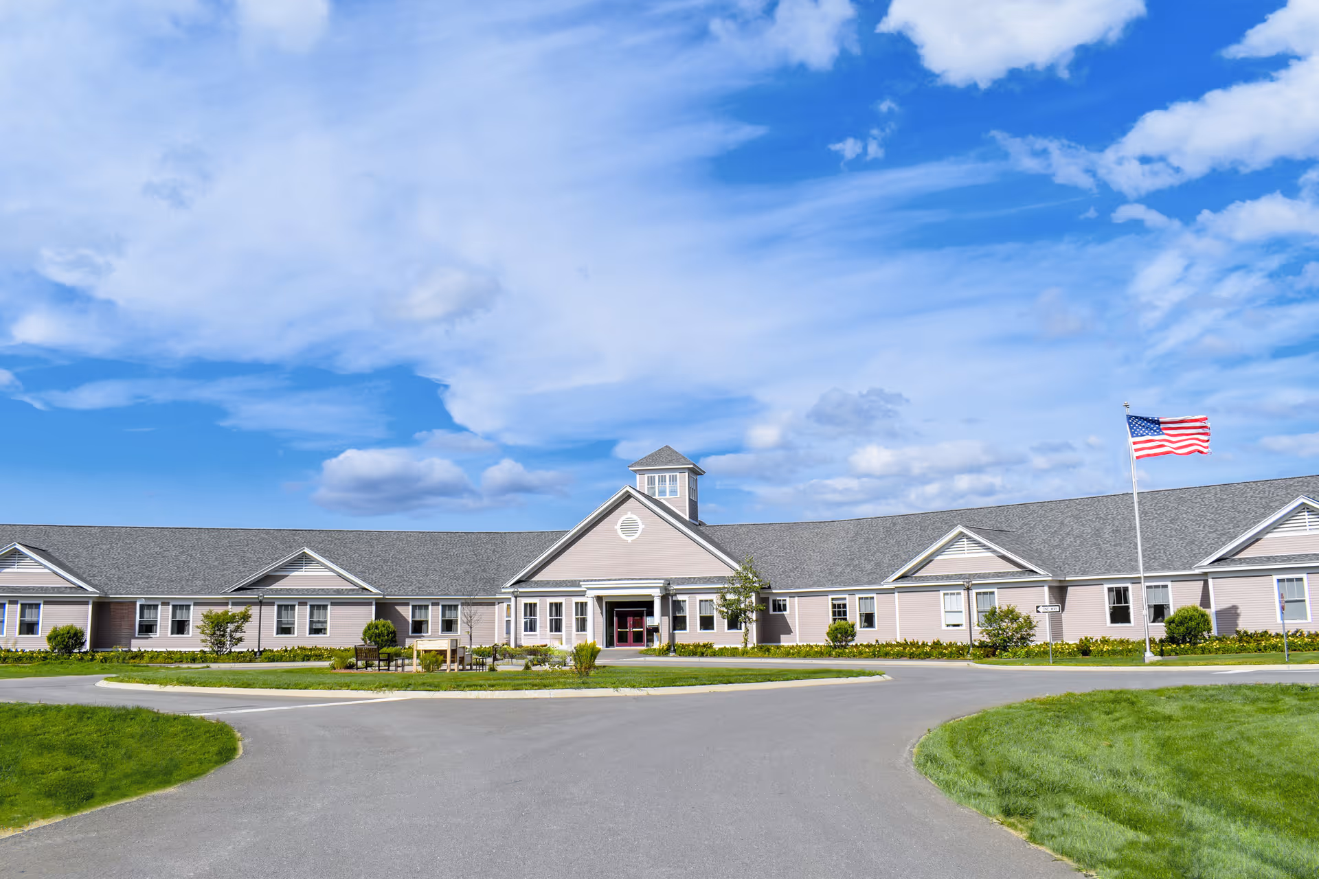 Front view of a single-story senior living facility with a circular driveway, landscaped lawn, and an American flag under a blue sky.