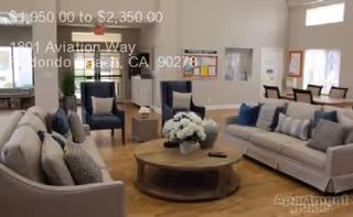 A spacious living room with two beige sofas and two dark blue armchairs arranged around a round wooden coffee table with a flower arrangement. The room has light-colored walls, large windows with white blinds, and wooden flooring. There is a fireplace in the corner and framed artwork on the walls.