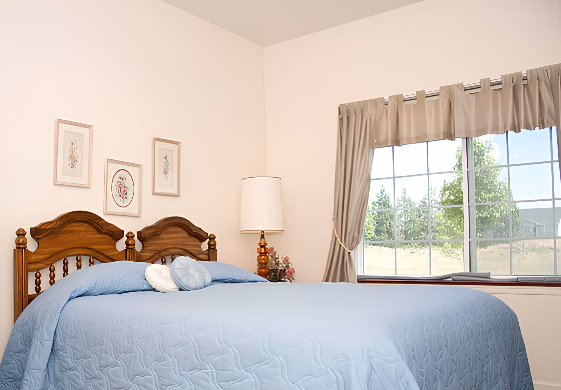 A bedroom with a large bed covered in a light blue quilt and two decorative pillows. The bed has a wooden headboard with a classic design. On the wall above the bed are three framed floral artworks. To the right of the bed is a wooden nightstand with a tall lamp and a small floral arrangement. A window with beige curtains tied back reveals a view of trees and a building outside.