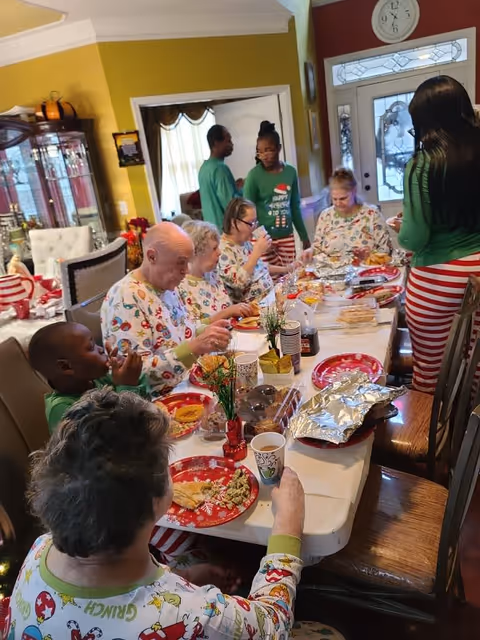 A group of elderly people and caregivers gathered around a dining table enjoying a meal together. The elderly individuals are wearing festive holiday pajamas with Christmas-themed prints, and some caregivers are wearing green tops and red-and-white striped pants. The table is set with red holiday plates, cups, and various food items. The setting appears to be a cozy dining room with warm yellow walls and a glass cabinet in the background.