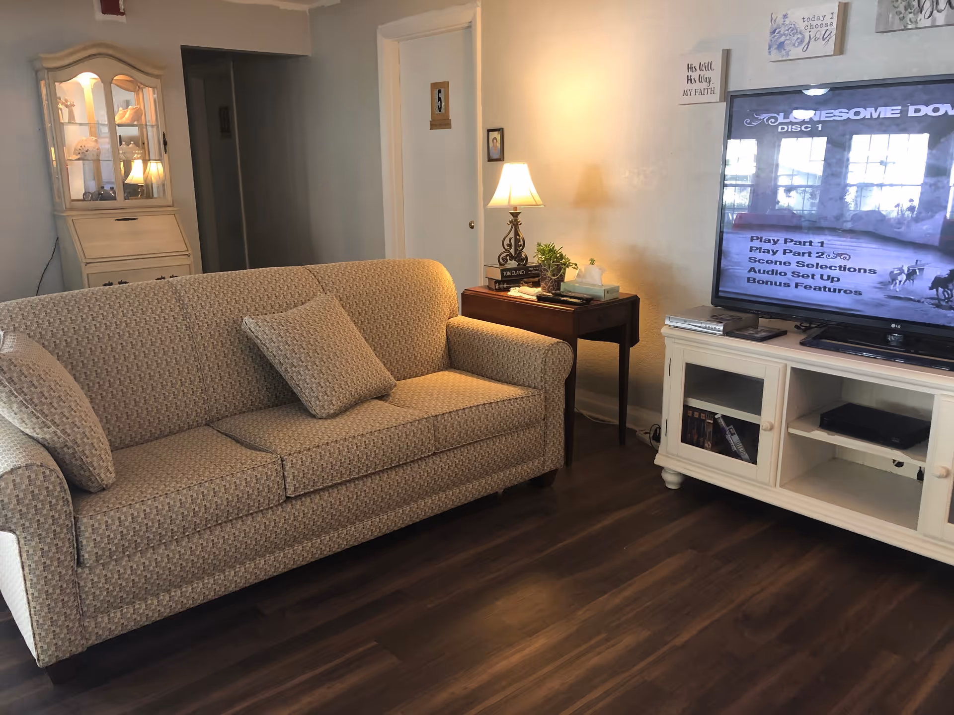 A cozy living room with a patterned beige sofa, a wooden side table with a lamp, books, and a plant, a white TV stand holding a flat-screen TV displaying a menu screen, and a glass-front cabinet with decorative items in the background.