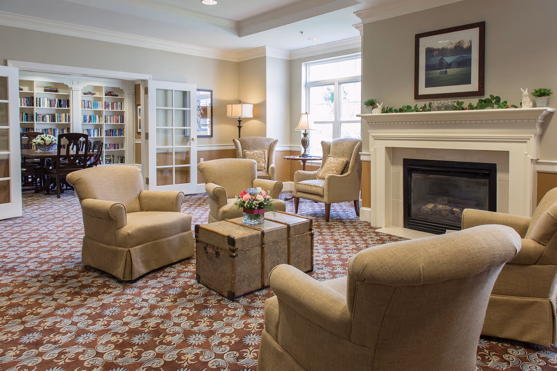 Cozy common living room with upholstered armchairs arranged around a trunk-style coffee table, a fireplace, and a book-filled room visible through glass double doors.