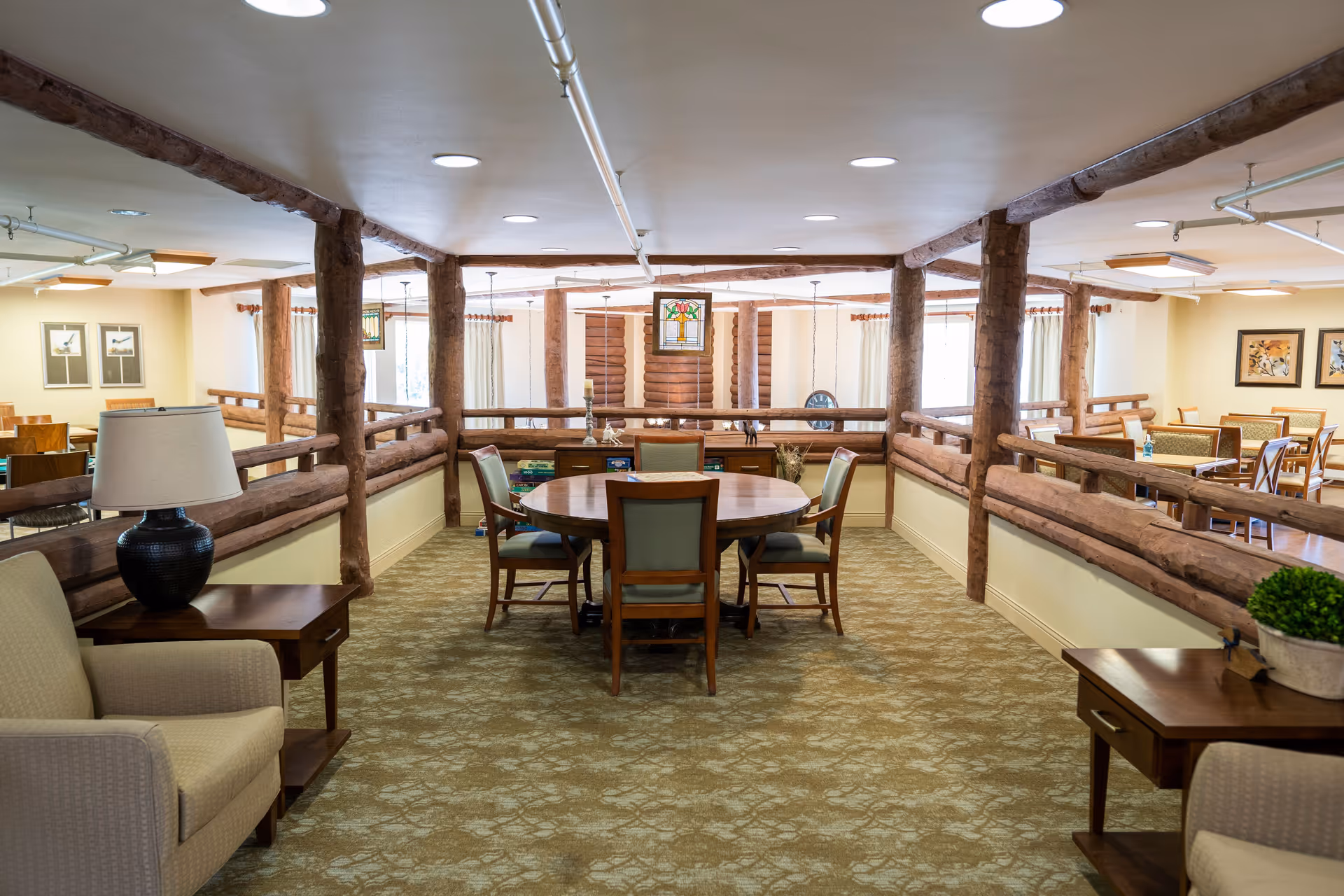 Interior view of a senior living facility common area with wooden log-style railings, a round wooden table with four chairs in the center, beige carpet with a floral pattern, armchairs with side tables and lamps, and framed artwork on the walls. The space is well-lit with ceiling lights and natural light from windows with curtains.