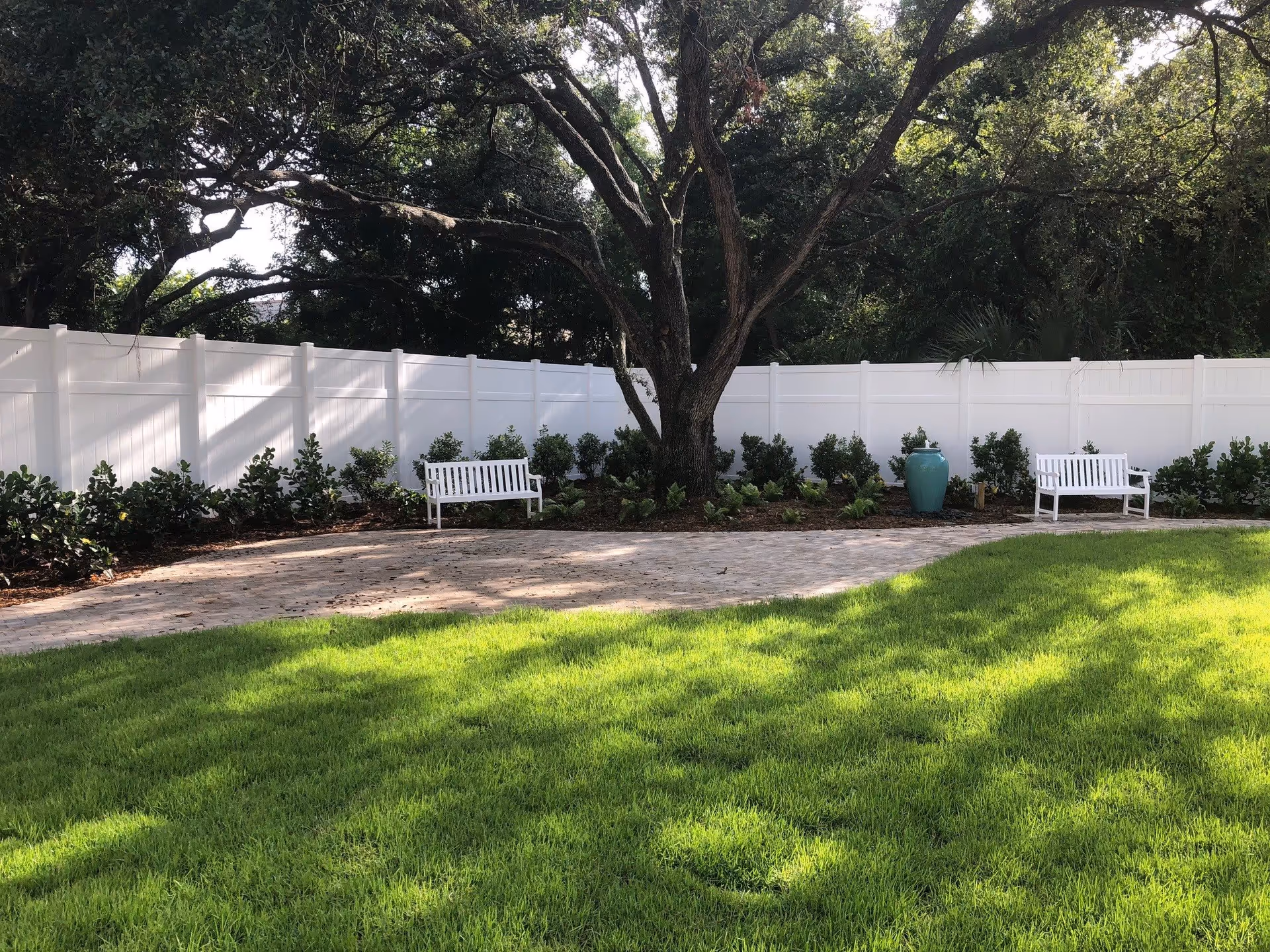 A peaceful outdoor garden area with a large tree in the center, surrounded by a white fence. There are two white benches on either side of the tree and a large blue ceramic pot near one bench. The ground is covered with green grass and a paved walkway.