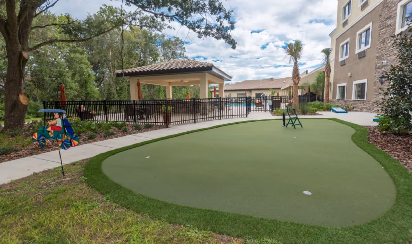 Outdoor area of a senior living facility featuring a putting green with two holes, a colorful decorative wind spinner shaped like a person on a bicycle, a covered patio with seating, palm trees, and a multi-story building with stone and stucco exterior under a partly cloudy sky.