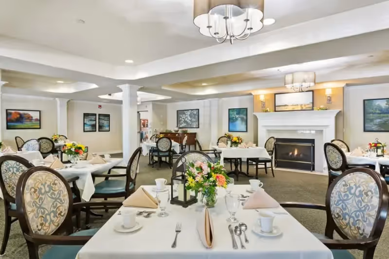 A bright and elegant dining room with several round tables covered with white tablecloths, each set with plates, cups, silverware, and floral centerpieces. The room features upholstered chairs with patterned backs, a fireplace with a painting above it, and soft overhead lighting.