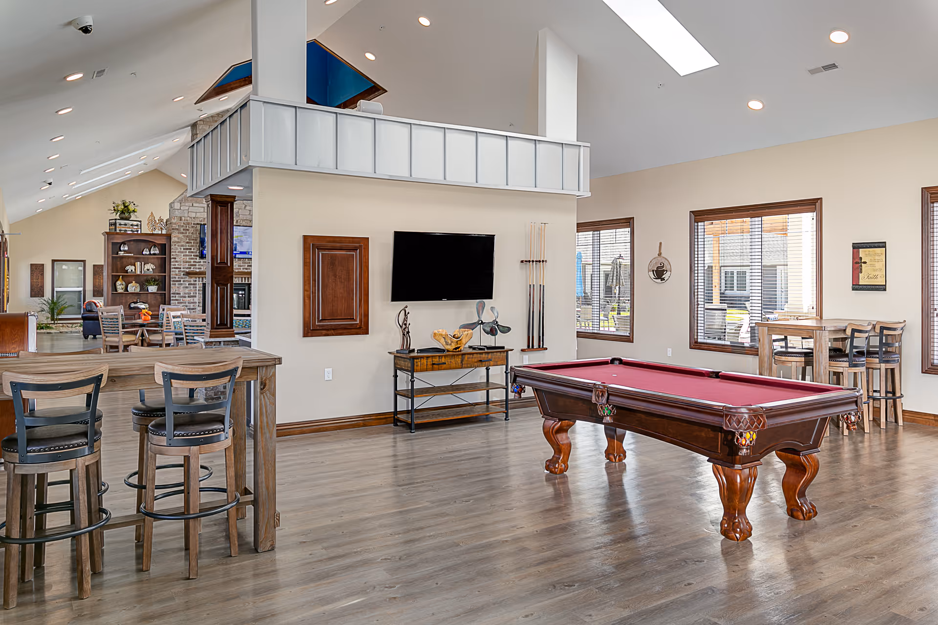 A spacious recreational room with a pool table featuring a red felt top in the center. The room has wooden flooring, multiple windows with blinds, and a wall-mounted TV above a small console table. There are several seating areas including high tables with chairs and a dining table in the background. The ceiling is vaulted with recessed lighting and skylights, and the walls are painted a light beige color.