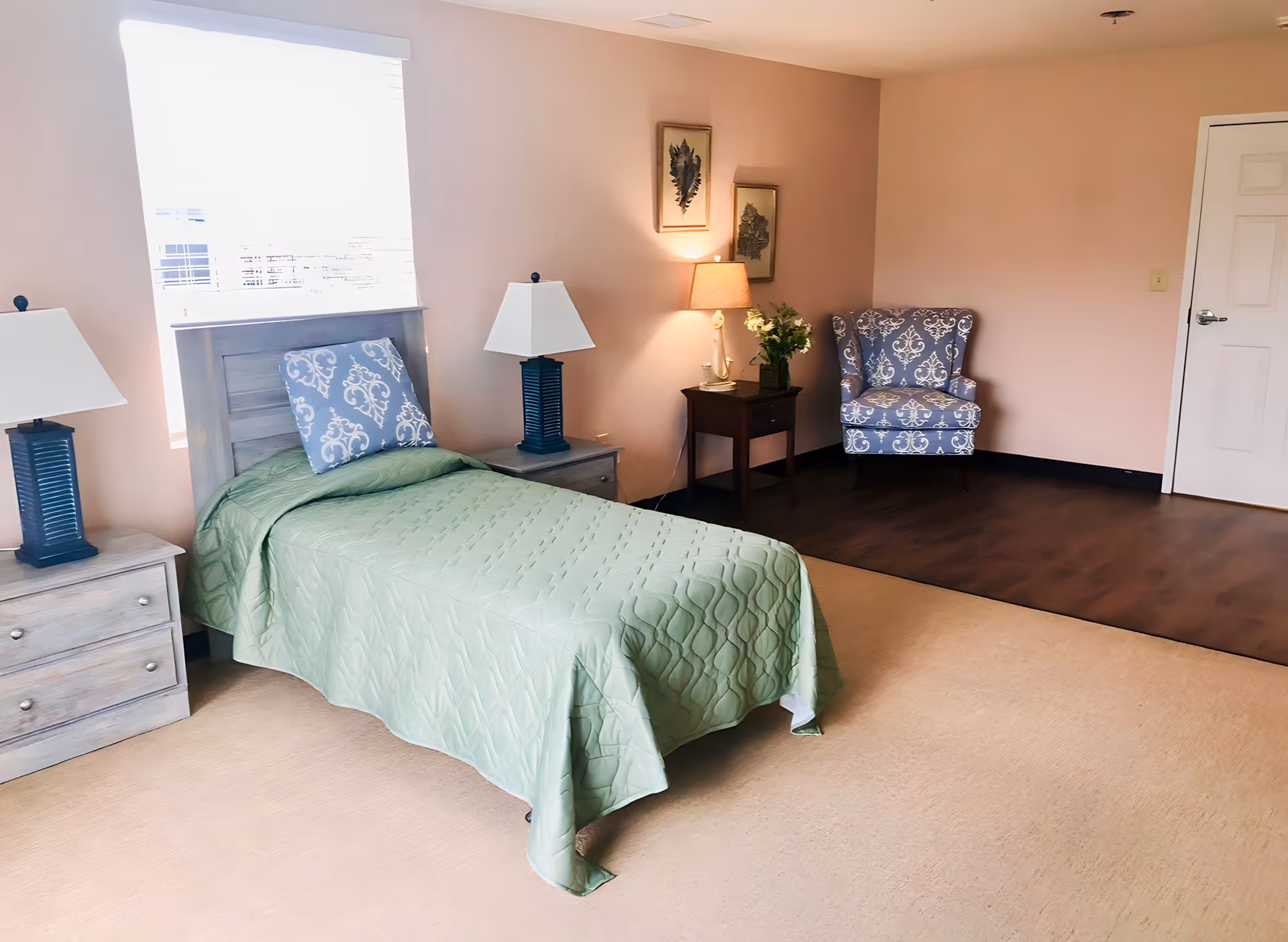Furnished single bedroom with a green-covered twin bed, two nightstands with lamps, and a patterned armchair in the corner.