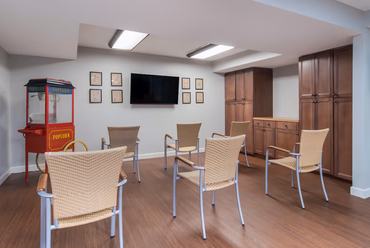 A small media room with six wicker chairs arranged in two rows facing a wall-mounted flat screen TV. On the left side of the room, there is a vintage-style red popcorn machine. The walls are light gray, and there are six framed pictures arranged around the TV. On the right side, there are tall wooden cabinets with a countertop. The floor is wood, and the ceiling has recessed lighting panels.