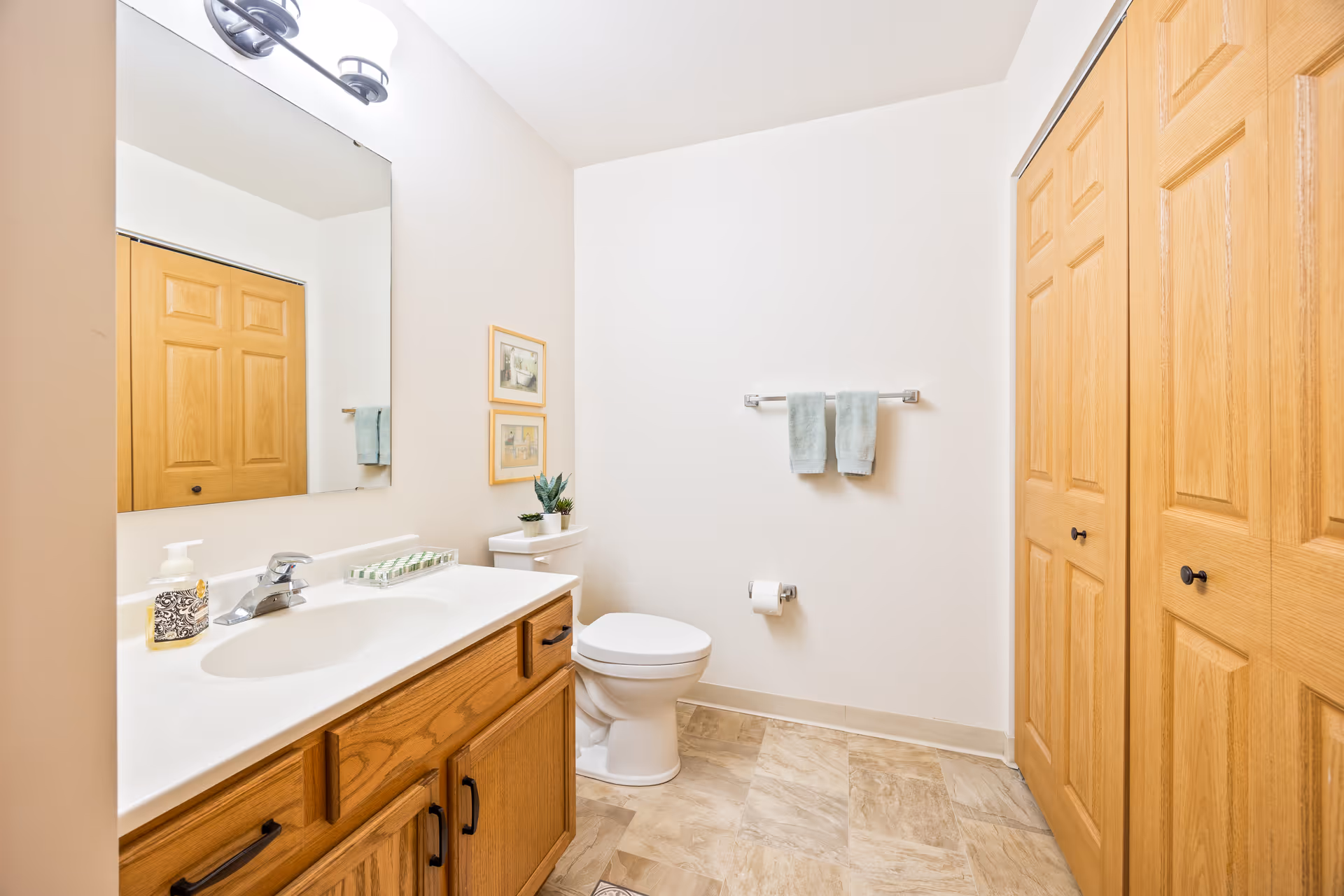 Bright bathroom with a wooden vanity and sink, mirror, toilet, towel rack, and double wooden closet doors.