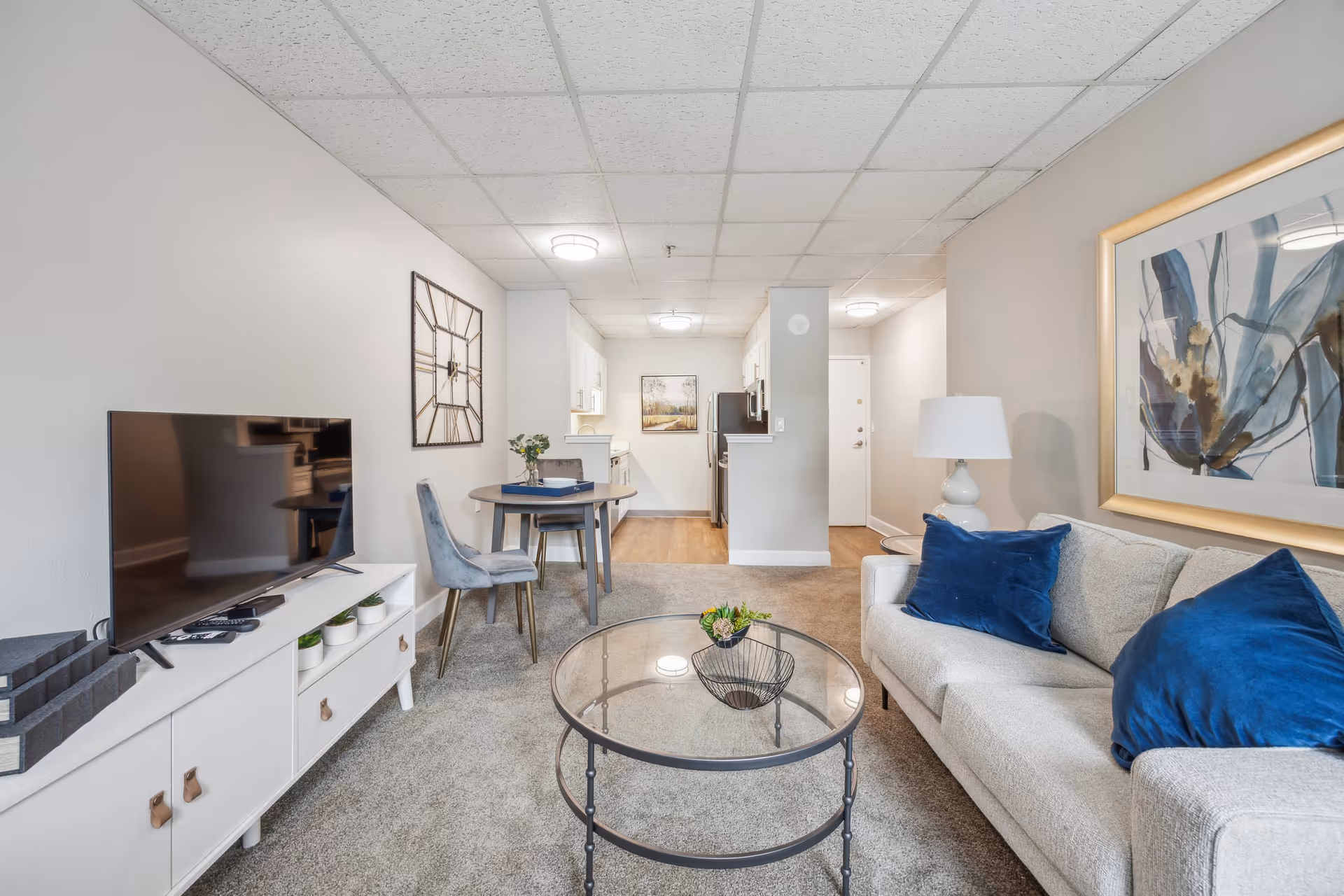 Bright living room with a sofa and blue pillows, glass coffee table, TV on a console, and a small dining area leading to a kitchenette.
